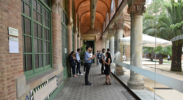 People talking to each other a hall filled with natural light. Outside, a palm tree offers shade.